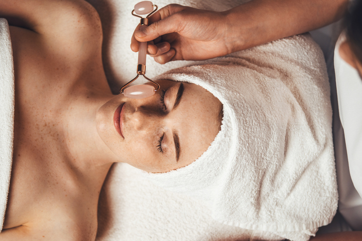 Close up portrait of beautician's hand massaging patient's face with jade roller at spa salon. Beauty woman face skin care. Body care, spa.